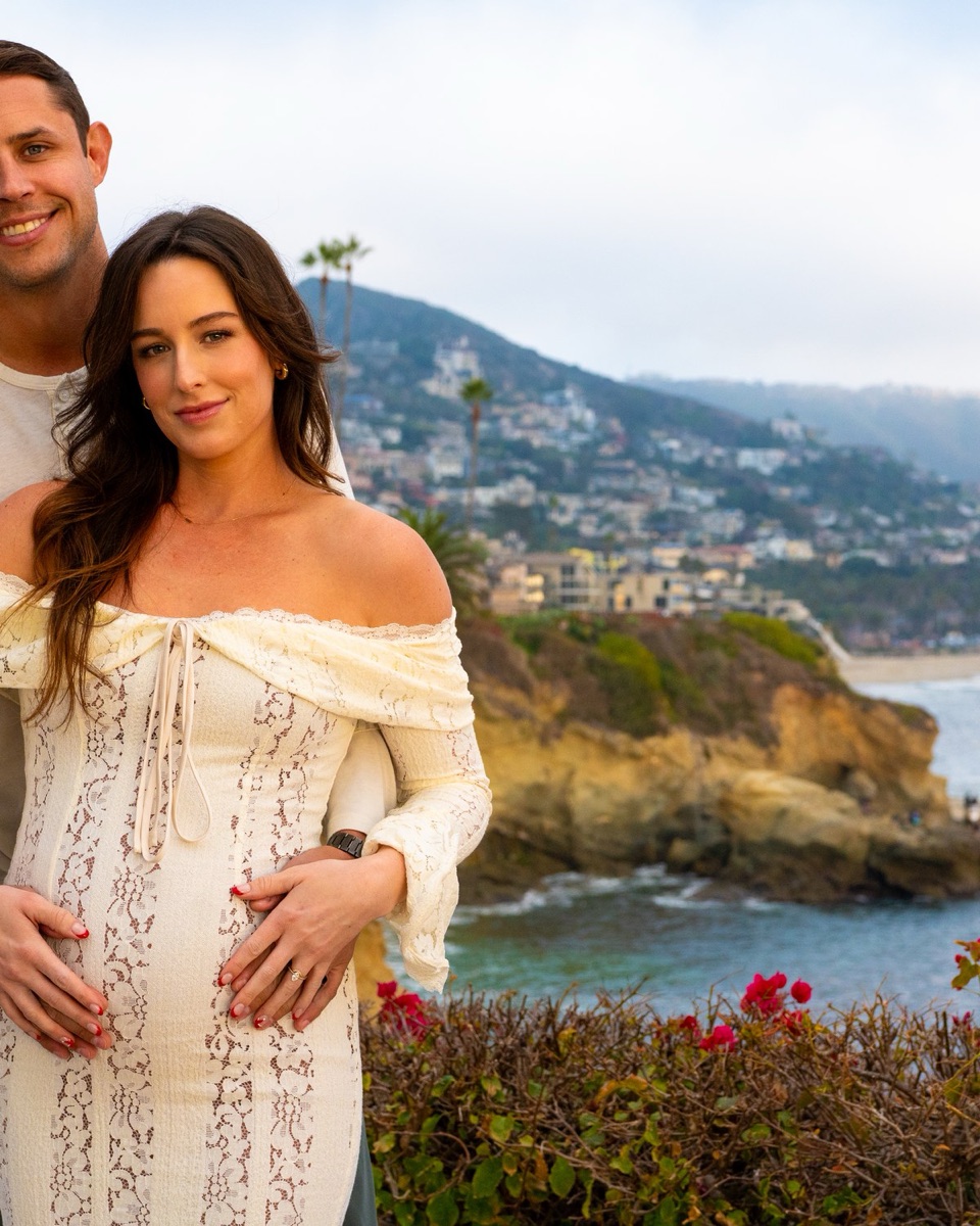 Pregnant woman leaning back against her partner on a coastal bluff, his hands resting on her belly