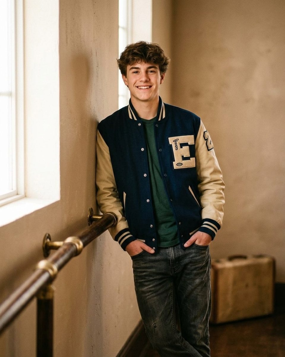 Senior in a varsity jacket leaning back against a wall beside a window, hands in his pockets