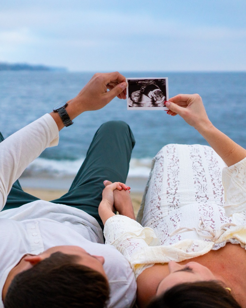Couple on a beach at dusk holding a sonogram print up together, looking at it