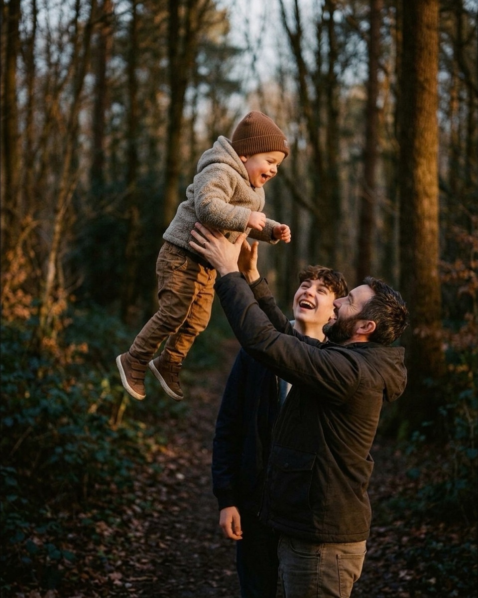 Father tossing a toddler into the air on a forest path with a teen looking on, backlit by low fall sun
